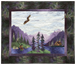 Quilt block showing a fisherman standing in a river casting a fly, with an eagle flying overhead. The riverside cabin and large purple mountains are in the background.