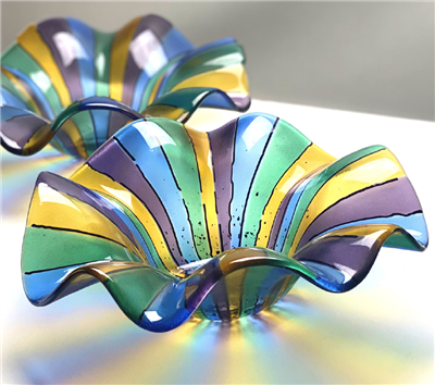Two glass bowls on a white table with light shining through them. Each bowl has yellow, green, blue, and purple stripes starting from the center with thin black lines between each color and a few flecks of black in the center. The bowl edges are wavy.