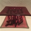 An artistic sculpture made of burgundy glass. The top is a square with several holes in the middle, and the base is formed from curving strings of glass that dripped through these holes as the glass heated. Light shines through onto the white table below.
