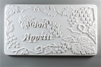 A long rectangular tile made of white ceramic. The carved texture on it reads Bon Appetit in script just to the left of center. The words are surrounded by bunches of grapes and leaves on vines.