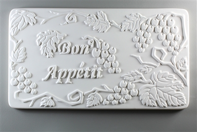 A long rectangular tile made of white ceramic. The carved texture on it reads Bon Appetit in script just to the left of center. The words are surrounded by bunches of grapes and leaves on vines.