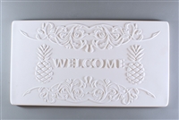 A long rectangular tile made of white ceramic. The carved texture says Welcome in the center. The word is flanked on either side by a pineapple, and the bottom and top of the tile have elaborate curving lines drawing attention inwards to the word.