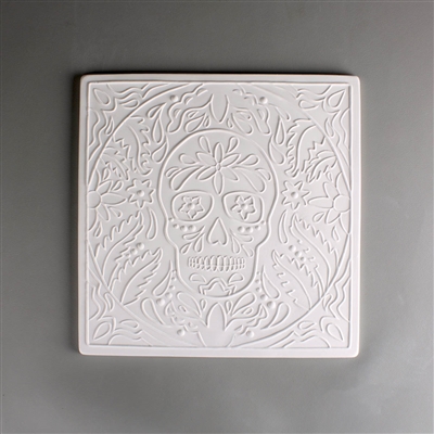 A large square tile made of white ceramic. The carved texture shows a human skull in the center decorated in the style of the Day of the Dead. A circle of similar designs surrounds the skull with a square of simpler designs around it.