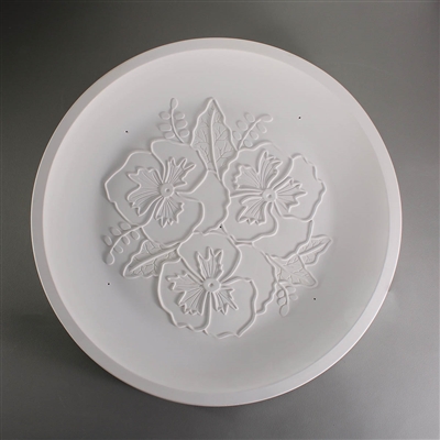 A circular white ceramic mold for fusing glass on a grey background. The edge is raised slightly to create a dish shape. The flat center has been carved with a group of three pansy flowers surrounded by leaves and small vines.