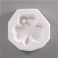 A roughly circular, white ceramic mold for fusing hot glass on a grey background. A three-leaved shamrock clover has been carved into it. There is a small post on the left side of the top leaf of the clover so the finished piece can be strung as jewelry.