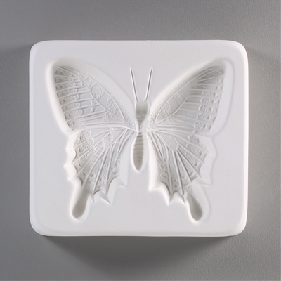 A square, white ceramic mold for fusing hot glass on a grey background. A very detailed flat swallowtail butterfly has been carved into it.