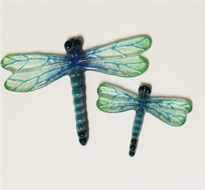 Two fused glass dragonflies on a cream background. Each dragonfly has an opaque teal and black body and clear transparent wings with light blue veins and greenish tips. The left dragonfly is slightly larger. Both dragonflies face upwards.