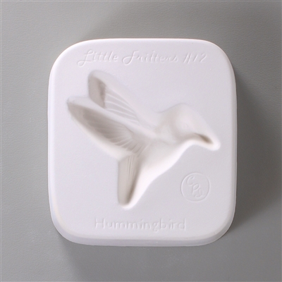 A square, white ceramic mold for fusing hot glass on a grey background. A simple profile of a hummingbird in flight has been carved into it. The mold has Little Fritters #12 Hummingbird engraved on the sides in script.