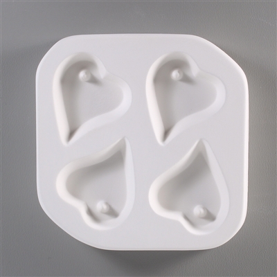 A square, white ceramic mold for fusing hot glass on a grey background. Four small hearts with curved tips have been carved into it with equal distance between them. Each heart has a post in its top left to allow for stringing into jewelry after firing.