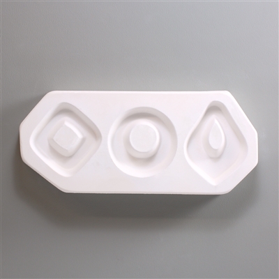 A rectangular, white ceramic mold for fusing hot glass on a grey background. Three large geometric shapes have been carved into it, each with uncarved middles for stringing into jewelry. The left is a diamond, the middle a circle, and the right a tear.