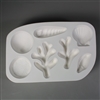 A rectangular white ceramic mold for fusing hot glass on a grey background. Four different kinds of shell, one sand dollar, and two pieces of branching coral have been carved into it. Each piece has a slightly different design, but all are similarly sized