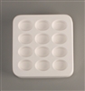 A square white ceramic mold for fusing hot glass on a grey background. Twelve identical ovals have been carved into it with equal space between them.