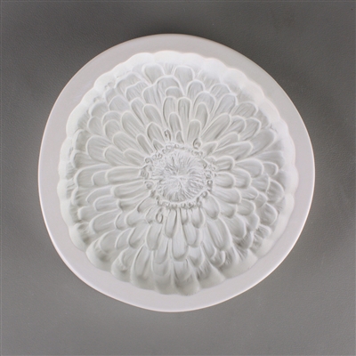 A circular, white ceramic mold for fusing hot glass on a grey background. An intricately detailed flat zinnia flower has been carved into it. The flower takes up most of the mold, but there is a small border of empty space around it.