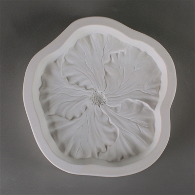 A roughly circular, white ceramic mold for fusing hot glass on a grey background. A detailed flat hibiscus flower has been carved into it. The flower takes up most of the mold, but there is a small border of empty space around it.
