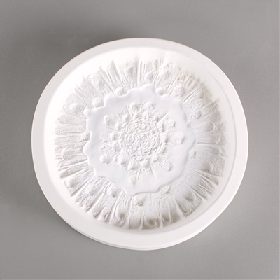 A circular white ceramic mold for fusing hot glass on a grey background. A detailed but flattened mushroom cap has been carved into it. The cap takes up most of the mold, but there is a small border of empty space around it.