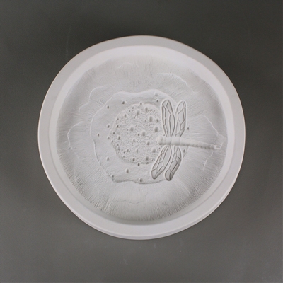 A circular white ceramic mold for fusing hot glass on a grey background. A detailed but flattened mushroom cap with a dragonfly on its center has been carved into it. The cap takes up most of the mold, but there is a small border of empty space around it.