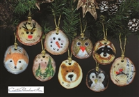 Nine different fused glass ornaments hang from an evergreen branch in front of a dark background. Each ornament is strung with gold thread and has a base that looks like a log slice. Each ornament has a different design such as a snowman or deer face.