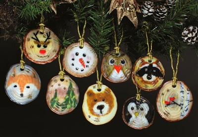 Nine different fused glass ornaments hang from an evergreen branch in front of a dark background. Each ornament is strung with gold thread and has a base that looks like a log slice. Each ornament has a different design such as a snowman or deer face.