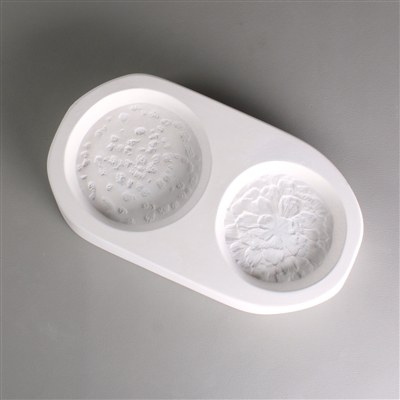 An oval-shaped white ceramic mold for fusing hot glass on a grey background. Two detailed flat mushroom caps have been carved into it. The left cap is slightly larger.