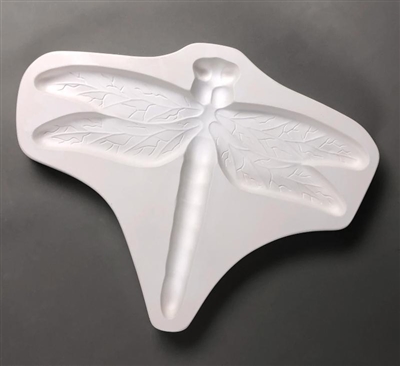 A triangular white ceramic mold for fusing hot glass on a grey background. A large and detailed dragonfly has been carved into it.