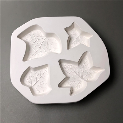 A circular white ceramic mold for fusing hot glass on a grey background. Four ivy leaves have been carved into it. Each leaf is shaped and sized slightly differently, with two having three points and two having five.