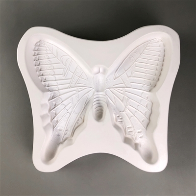 A large white ceramic mold for fusing hot glass on a grey background. A large and intricately detailed butterfly has been carved into it.