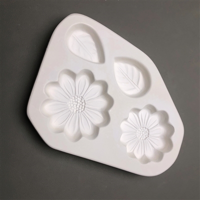 A roughly triangular white ceramic mold for fusing hot glass on a grey background. Two small daisy flowers and two teardrop leaves have been carved into it. The daisies are different sizes with similar designs, and the leaves are about the same.