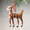 A fused glass standing deer ornament hanging from a pine branch on a grey background by a red string. The deer is mostly brown with a tan face, chest, and lower legs. It is wearing a red harness on its chest with gold jingle bells and has black hooves.