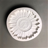 A circular white ceramic mold for fusing hot glass on a grey background. A detailed flat sunflower has been carved into it. The flower takes up most of the mold, but there is a small border of empty space around it.