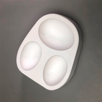 A roughly oval white ceramic mold for fusing hot glass on a grey background. Three separate large eggs in two different sizes have been carved into it. The top egg is larger while the two eggs below it are smaller.