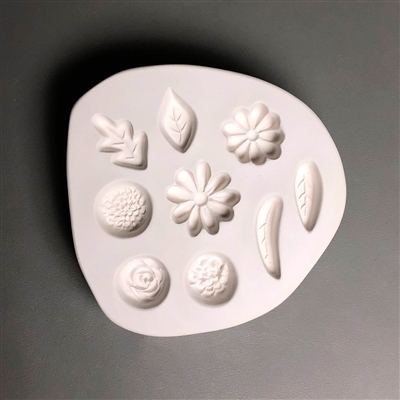 A roughly circular white ceramic mold for fusing hot glass on a grey background. An assortment of small flowers and leaves have been carved into it. There are two daisies, a rose, a mum, a zinnia, and four separate shapes of leaf.