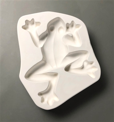 A white ceramic mold for fusing hot glass on a grey background. A top-down view of a large frog has been carved into it. The frog is plain in design and facing slightly to the right. Its arms are bent around either side of its head and its legs are bent.