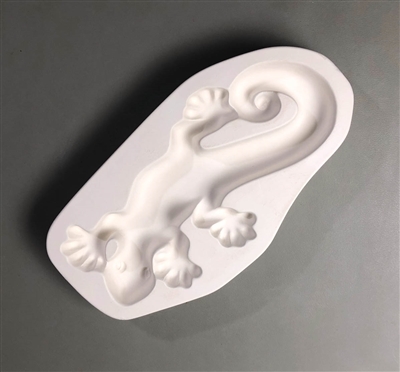 A roughly oval white ceramic mold for fusing hot glass on a grey background. A top-down view of a crawling lizard has been carved into it. The lizard is simple in design and facing downwards. The tip of its tail curls in on itself.