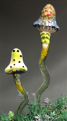 Two mushroom shaped garden stakes made of fused glass. The left one is shorter, with a lime green mushroom cap with black spots. The right cap is mostly grey with red and green details on top. The stakes fade from olive to light green.