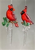 Two fused glass cardinal ornaments hang in front of a gray background. The cardinals are red with black markings and orange beaks. Each sits on a small twig of greenery that has long clear icicles hanging from it.