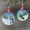 Two round fused glass ornaments hang from a branch. Each ornament shows a snowy landscape featuring a blue and white sky, white snow, and triangular green pine trees. Both have red tops and a few drawn silver snowflake decorations.