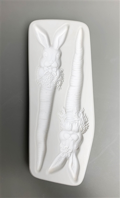 A rectangular white ceramic mold for fusing hot glass on a grey background. Two rabbit faces have been carved into it. The left rabbit face has a bow tie and the right has a flower on its ear. Both are positioned on top of long carrot stakes.