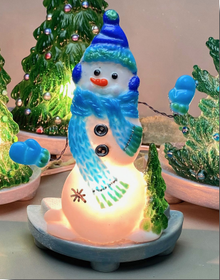 A fused glass snowman displayed on a blue base with a light behind it. The snowman has a hat, scarf, earmuffs, and mittens in various shades of blue and stands next to a small green pine tree. The mittens are attached with twisted wire.