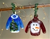 Two fused glass sweater ornaments hang from a branch with small lights wrapped around it. The left sweater is blue with white trim and a colorful Christmas tree design. The right sweater is red with painted gold trim and a large smiling snowman face.