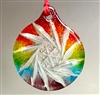 A round, fused glass Christmas ornament hanging from a red string in front of a grey background. The ornament has a transparent spiral cut crystal design with a rainbow gradient surrounding it, going from red at the top to purple at the bottom.