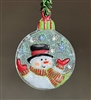 A round, fused glass ornament hanging in front of a grey background. The ornament has a smiling snowman on an iridescent clear background with silver and white snowflakes. He has a green and red striped scarf as does the top of the ornament.