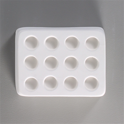 A rectangular white ceramic mold for fusing hot glass on a grey background. Twelve small separate but identical circles have been carved into it with equal space between them.