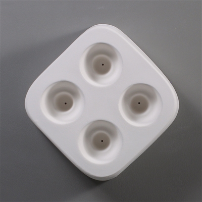 A square white ceramic mold for fusing hot glass on a grey background. Four separate identical circles have been carved into it with equal space between them. Each circle has a deeper circle carved in its center with a small vent hole in the center.