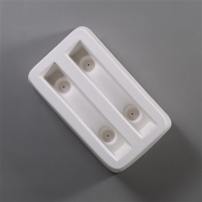 A rectangular white ceramic mold for fusing hot glass on a grey background. Two separate identical rectangles have been carved into it. Each rectangle has two deeper circles carved towards each end with a small vent hole in the center of each.