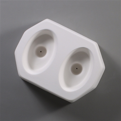 A white ceramic mold for fusing hot glass on a grey background. Two separate identical ovals have been carved into it with a space between them. Each oval has a deeper circle in the center with a small vent hole in the very center.