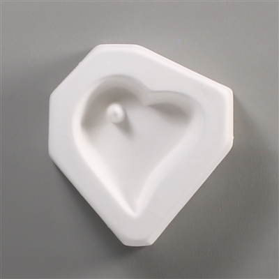 A white ceramic mold for fusing hot glass on a grey background. A slightly curved heart has been carved into it. There is a post towards the top left of the heart allowing for the resulting glass to be strung as jewelry.