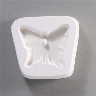 A trapezoidal white ceramic mold for fusing hot glass on a grey background. A simple butterfly has been carved into it. There is a post towards the top of the butterflyâ€™s head allowing for the resulting glass to be strung as jewelry.