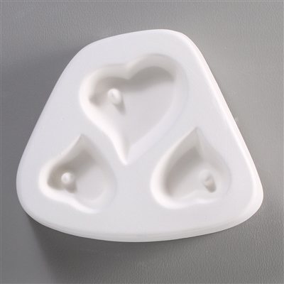 A white ceramic mold for fusing hot glass on a grey background. Three hearts have been carved into it, one large at the top and two smaller below it on either side. There is a post towards the top left of each allowing for the resulting piece to be strung