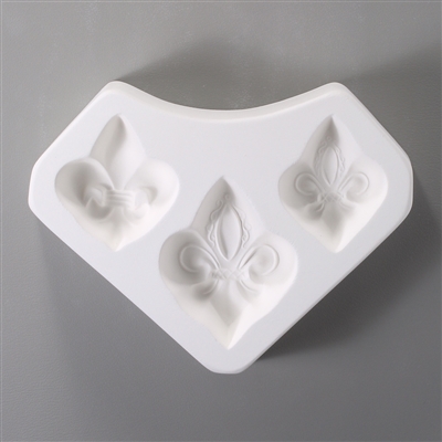 A white ceramic mold for fusing hot glass on a grey background. Three separate fleur de lis shapes have been carved into it. Each has slightly different detailing, and the one in the middle is marginally larger than the two on the sides.