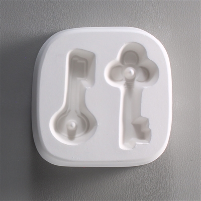 A square white ceramic mold for fusing hot glass on a grey background. Two different key shapes have been carved into it. The left key has a circular top and the right has a clover-shaped top. Both tops have posts to string the final glass pieces through.